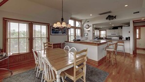 A spacious kitchen with a central island, wooden dining table with chairs, and large windows with blinds.