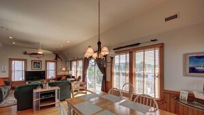 A spacious living room with a large TV, comfortable seating, a dining area with a wooden table and chairs, and large windows with blinds.