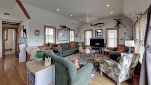 A spacious living room with a large sofa, a coffee table, and a television. There are several windows with wooden frames, and a large water dispenser is visible.