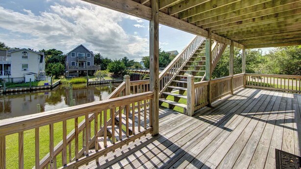 A wooden deck with a staircase leading to a covered area, overlooking a body of water and houses.