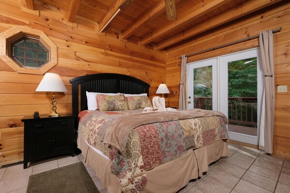 Quarto de cabana de madeira com cama, mesas de cabeceira e porta de correr para uma varanda.