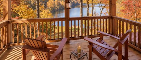 A wooden deck with Adirondack chairs and a small table, overlooking a lake and autumn foliage.