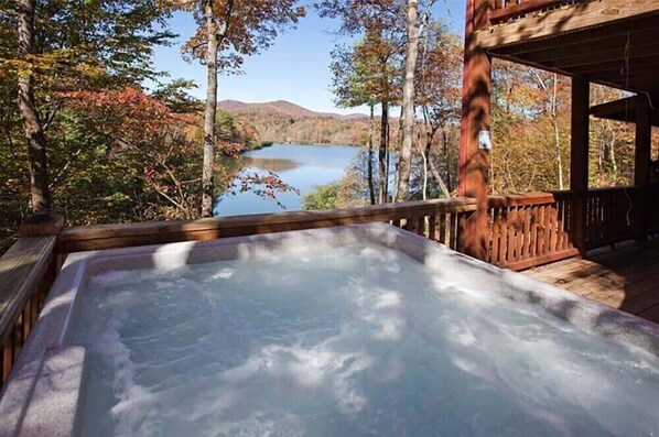 Relax in a hot tub on a deck overlooking a serene lake surrounded by trees with vibrant foliage.