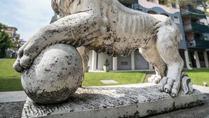 A stone lion statue with a ball, set on a pedestal, in front of a modern building with balconies.