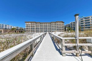 Une promenade en bois mène à un hôtel en bord de mer, doté de nombreux balcons, sous un ciel d’un bleu limpide.