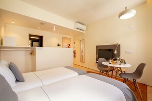 A modern hotel room with a bed, a dining table with chairs, a TV, and an air conditioning unit.