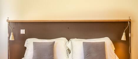 Cama doble con sábanas blancas y dos almohadas grises, cabecera de madera y dos lámparas de noche.