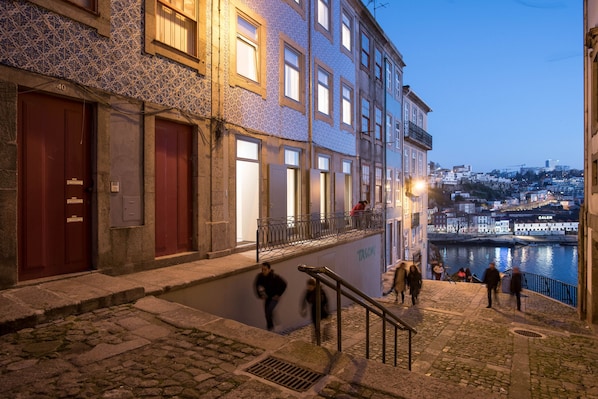 Une scène urbaine nocturne avec une rue pavée, des bâtiments aux façades ornées de motifs et une rivière en arrière-plan.