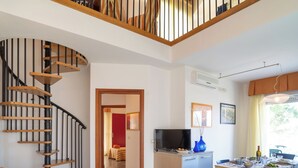 Amplia sala de estar con escalera de madera, comedor con mesa y sillas, y cocina con televisor y un jarrón azul.