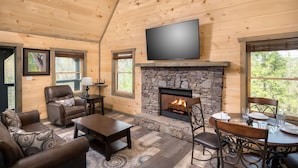 Interior acogedor de una cabaña con chimenea de piedra, paredes de madera, televisor de pantalla plana, sillón de cuero y comedor.
