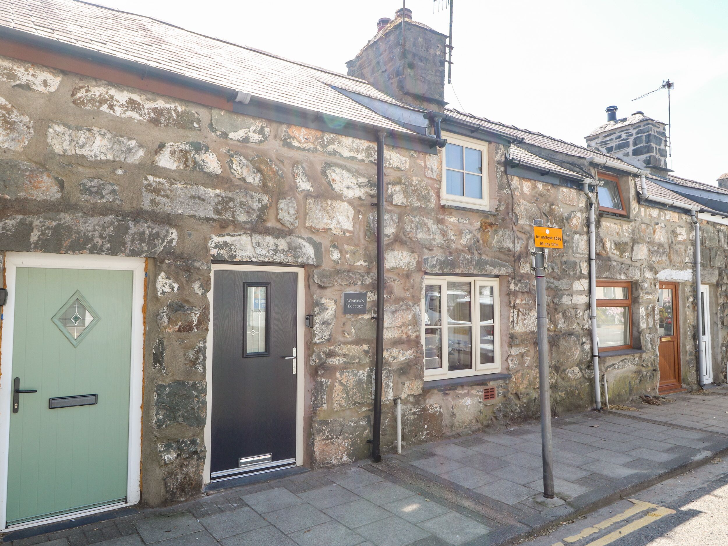 A stone building with a green door, a black door, and a window. There is a sign on the wall and a yellow sign on a pole.