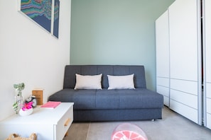 Un dormitorio moderno con un sofá gris, dos almohadas blancas, una mesita de noche blanca con un reloj, un libro rosa y una planta pequeña.