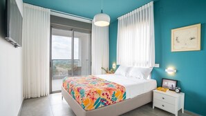 A bedroom with a bed, a nightstand, a window with curtains, a ceiling light, and a view of the outside. - Panoramic Sea Views Coastal Escape by Sea N' Rent (Nahariya)
