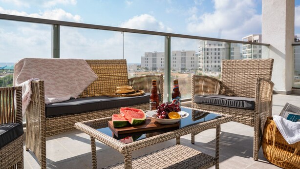 A balcony with wicker furniture, a table with food and drinks, and a view of buildings in the background. - Panoramic Sea Views Coastal Escape by Sea N' Rent (Nahariya)