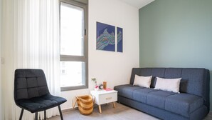 A modern living room with a grey sofa, a black chair, a white bedside table with a lamp, and a window with sheer curtains. - Panoramic Sea Views Coastal Escape by Sea N' Rent (Nahariya)