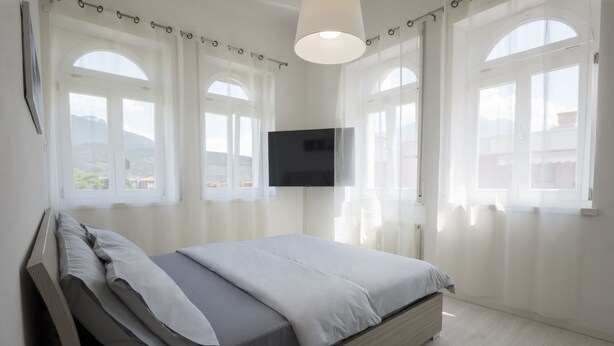 Una camera da letto con un letto, una televisione montata a parete e ampie finestre con tende.