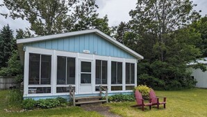 A small, single-story building with a blue exterior and white trim. It features large windows and a small porch area with two Adirondack chairs. - Charming cottage with shared beachfront on Grand Traverse Bay near Northport. (Northport)