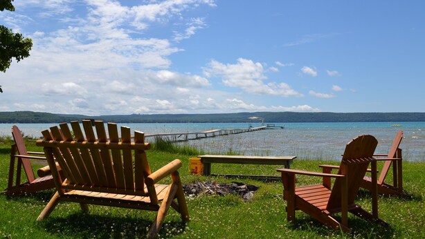 A lakeside setting with wooden Adirondack chairs and a picnic table, a dock extending into the water, and a clear sky.