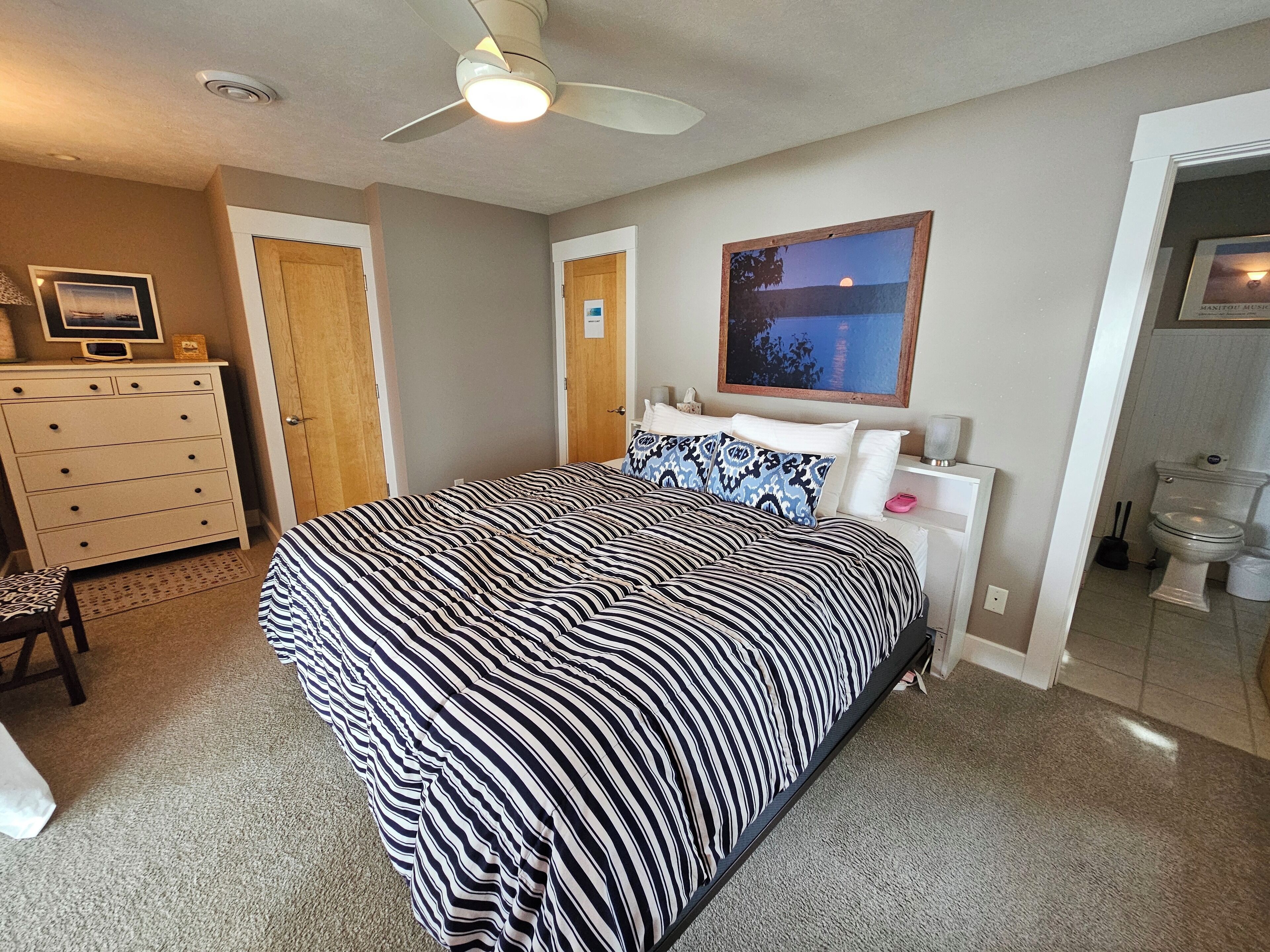 Une chambre à coucher avec un lit, une commode, un ventilateur de plafond, un tableau encadré et une salle de bain visible à travers une porte ouverte.