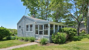 A small, single-story house with a covered porch, surrounded by lush greenery and a clear blue sky.