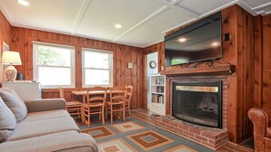 A cozy living room with a fireplace, wooden walls, a TV mounted above it, and a dining area with a table and chairs.