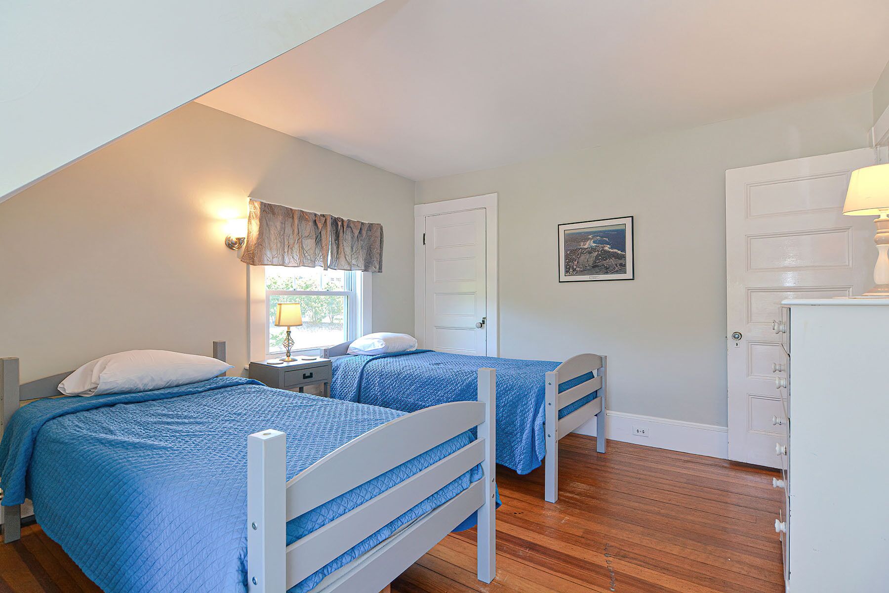 A bedroom with two beds, a window with curtains, a door, a wall-mounted picture, and a dresser.
