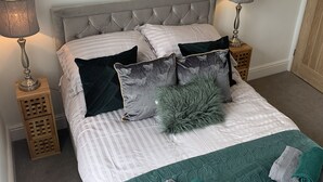 A neatly made bed with striped bedding, a tufted headboard, and decorative pillows. There are two bedside tables with lamps and a small wooden cabinet. - Beautiful 1-bed Apartment in Kirkby Lonsdale (Carnforth)