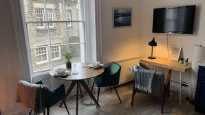 A cozy room with a round dining table, two chairs, a potted plant, a framed picture, a flat-screen TV, and a desk with a lamp. - Beautiful 1-bed Apartment in Kirkby Lonsdale (Carnforth)