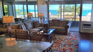A spacious living room with a large sofa, two armchairs, a coffee table, and a patterned rug. Large windows offer a view of the ocean and palm trees. - Kamaole Beach Royale 301 ~ 3 bedroom (Kihei)