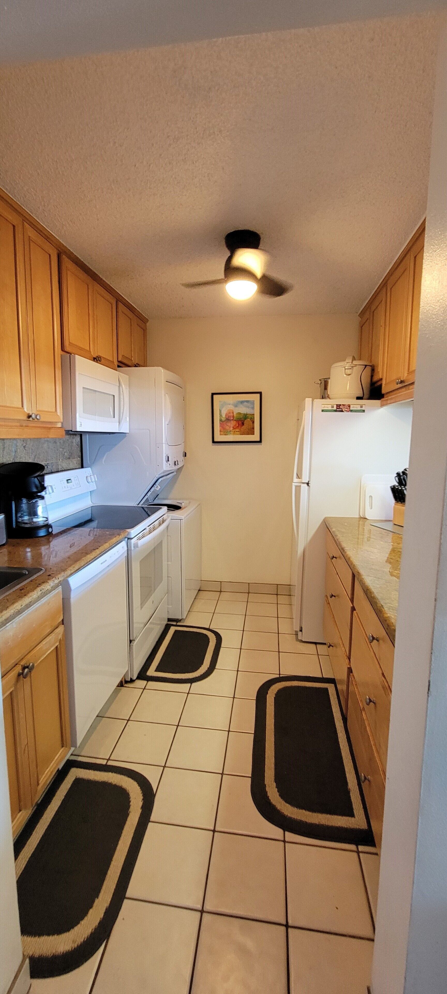 A compact kitchen with wooden cabinets, a microwave, a stove, and a refrigerator. There are two black and beige doormats on the tiled floor.