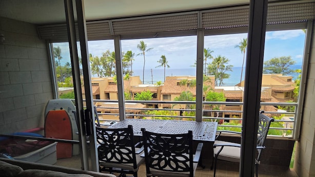 A balcony with a table and chairs, offering a view of palm trees and buildings across the water. - Kamaole Beach Royale 301 ~ 3 bedroom (Kihei)