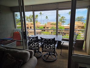 A balcony with a table and chairs, offering a view of palm trees and buildings across the water. - Kamaole Beach Royale 301 ~ 3 bedroom (Kihei)