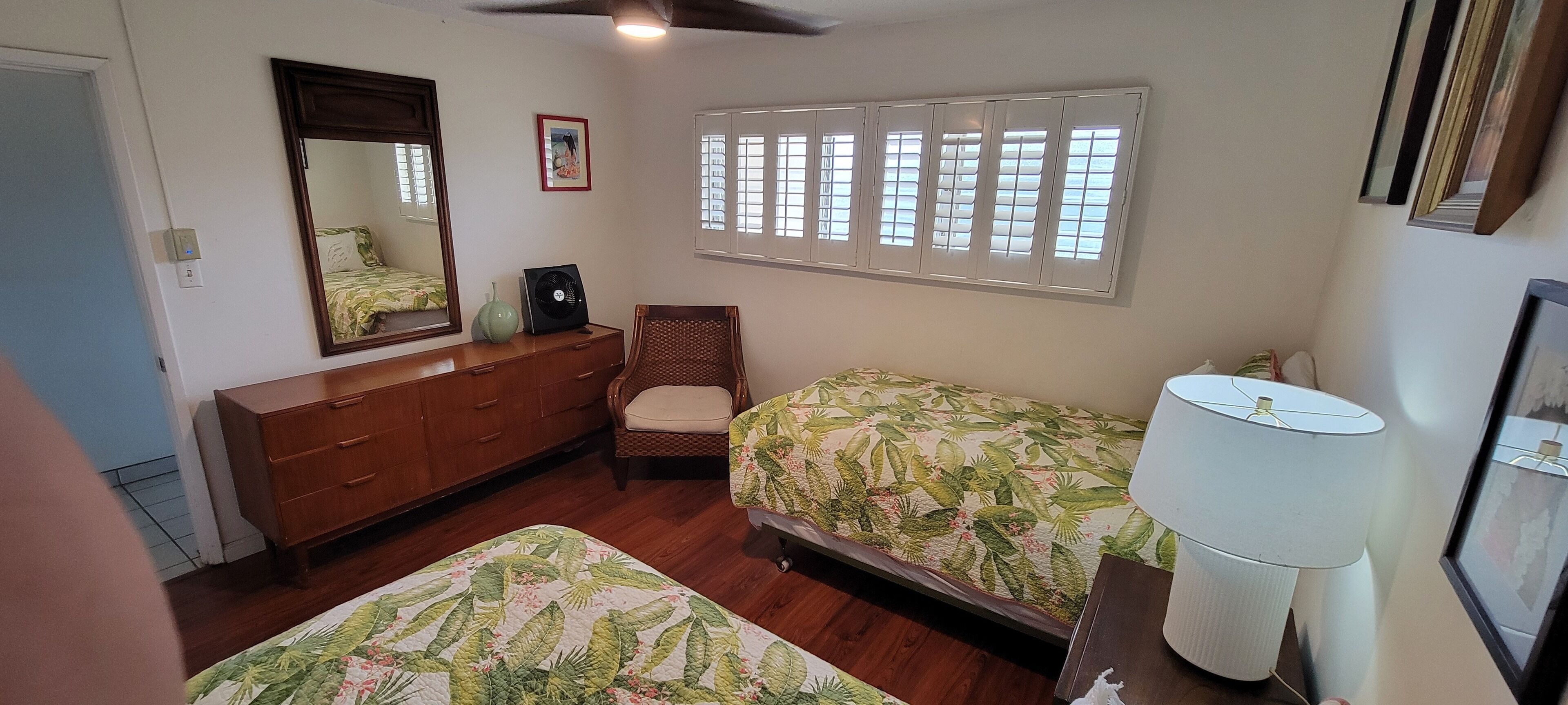 A bedroom with two beds, a dresser, a chair, a fan, and a window with shutters.