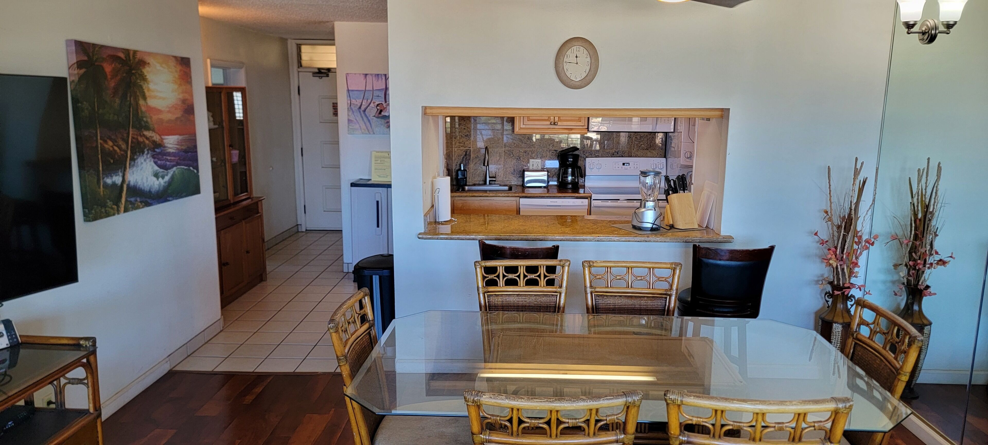 Een eethoek met een glazen tafel en rieten stoelen, een keuken met een ingebouwde oven en koffiezetapparaat, en een klok aan de muur.