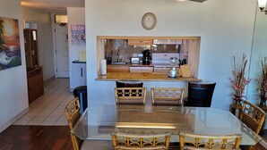 A dining area with a glass table and wicker chairs, a kitchen with a built-in oven and coffee maker, and a clock on the wall. - Kamaole Beach Royale 301 ~ 3 bedroom (Kihei)