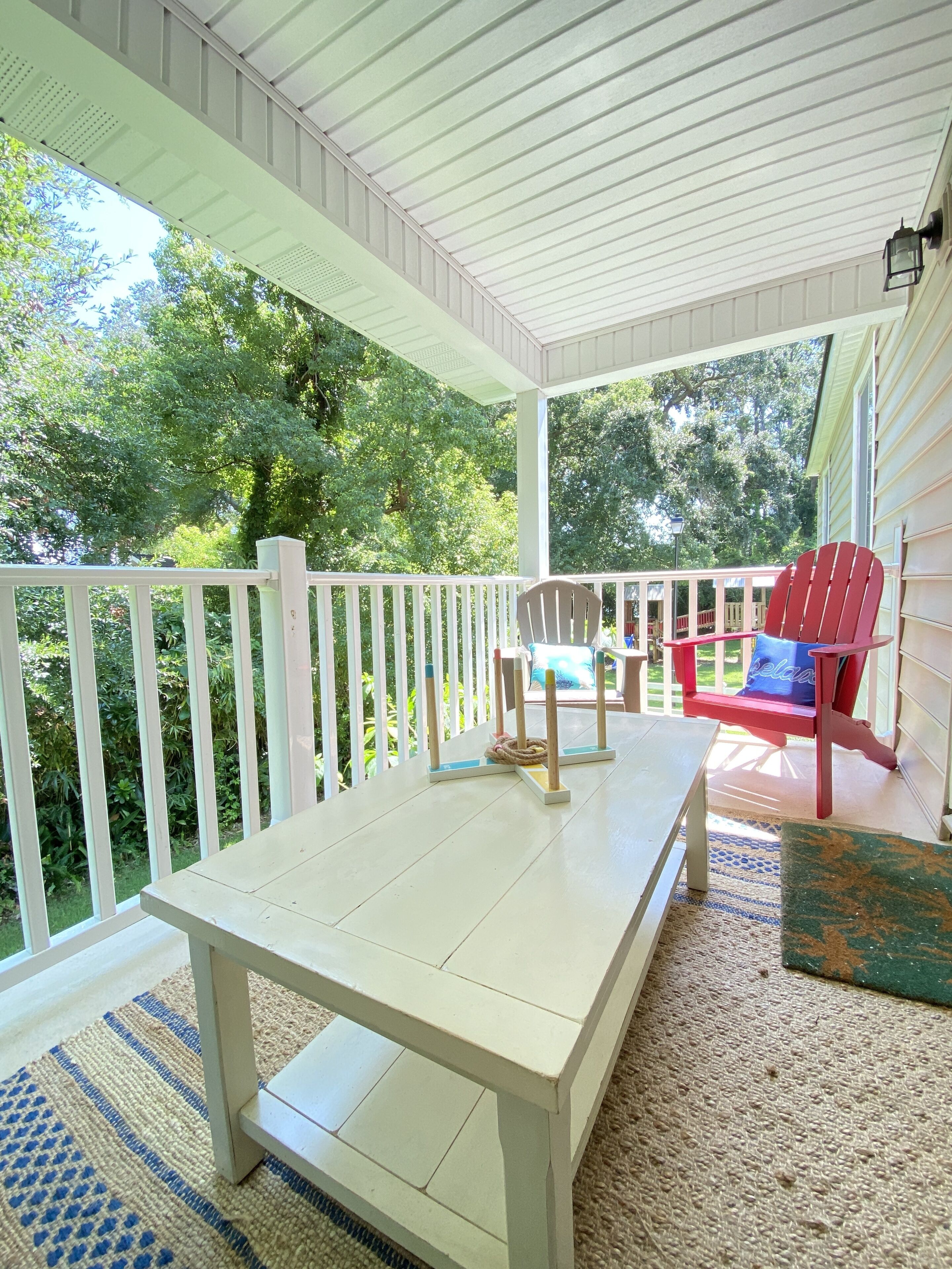 A covered porch with a white table and chairs, a red chair with a cushion, and a view of greenery.