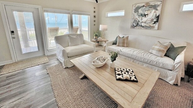 A spacious living room with a large wooden coffee table, two white sofas, and a view of the ocean through large sliding glass doors. - ALL DECKED OUT - 2 Bedroom, 2 Bath, Sleeps 6 - Beach Front! (Crystal Beach)