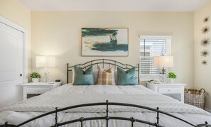 A bedroom with a bed, two bedside tables, a nightstand, a window with blinds, and a wall-mounted picture. - ALL DECKED OUT - 2 Bedroom, 2 Bath, Sleeps 6 - Beach Front! (Crystal Beach)