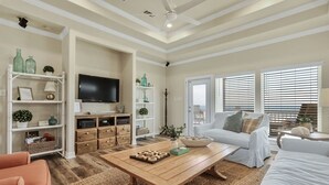 A living room with a white sofa, a wooden coffee table, and a TV on the wall. There are shelves with decorative items and a door leading to another room. - ALL DECKED OUT - 2 Bedroom, 2 Bath, Sleeps 6 - Beach Front! (Crystal Beach)