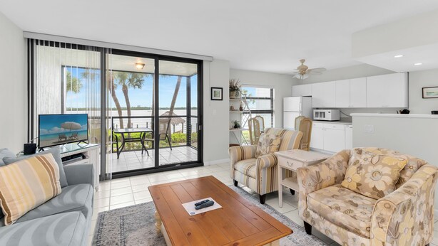 A modern living room with a sofa, a coffee table, and a TV. There is a sliding glass door leading to a balcony with outdoor furniture. - Riverside Club 1 Bed Overlooking Marco River (Marco Island)