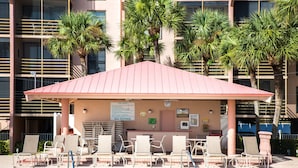 A pool area with lounge chairs and a covered seating area, surrounded by palm trees and a building with multiple balconies. - Riverside Club 1 Bed Overlooking Marco River (Marco Island)