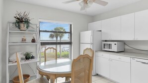 A kitchen with white cabinets, a microwave, and a dining area with a table and chairs. - Riverside Club 1 Bed Overlooking Marco River (Marco Island)