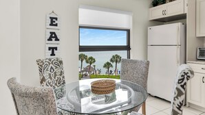 Een eetruimte met patroonstoelen, een glazen tafel, een bord met de tekst 'EAT', uitzicht op het strand en een keuken met een koelkast.