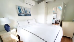 Un dormitorio con una cama grande, una mesita de noche con una lámpara, un cuadro con barcos de vela y vistas a la cocina a través de una puerta abierta.