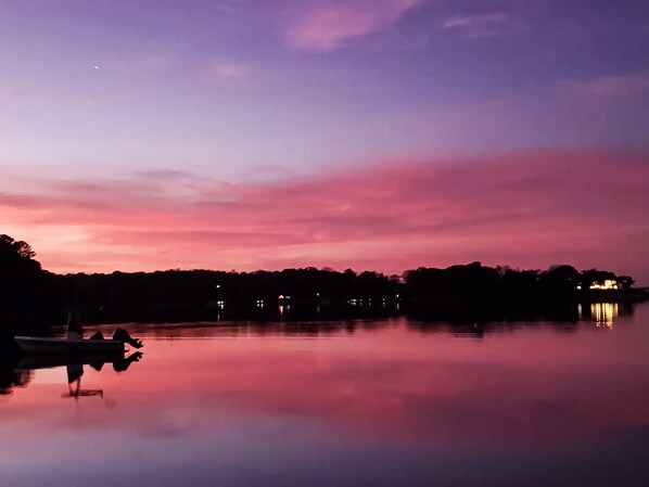 A calm lake at dusk with a boat docked on the left, a row of trees in the middle, and a building with lights on the right.