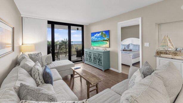 Een woonkamer met een bank, een salontafel en een televisie. Er is een balkon met uitzicht op de oceaan.