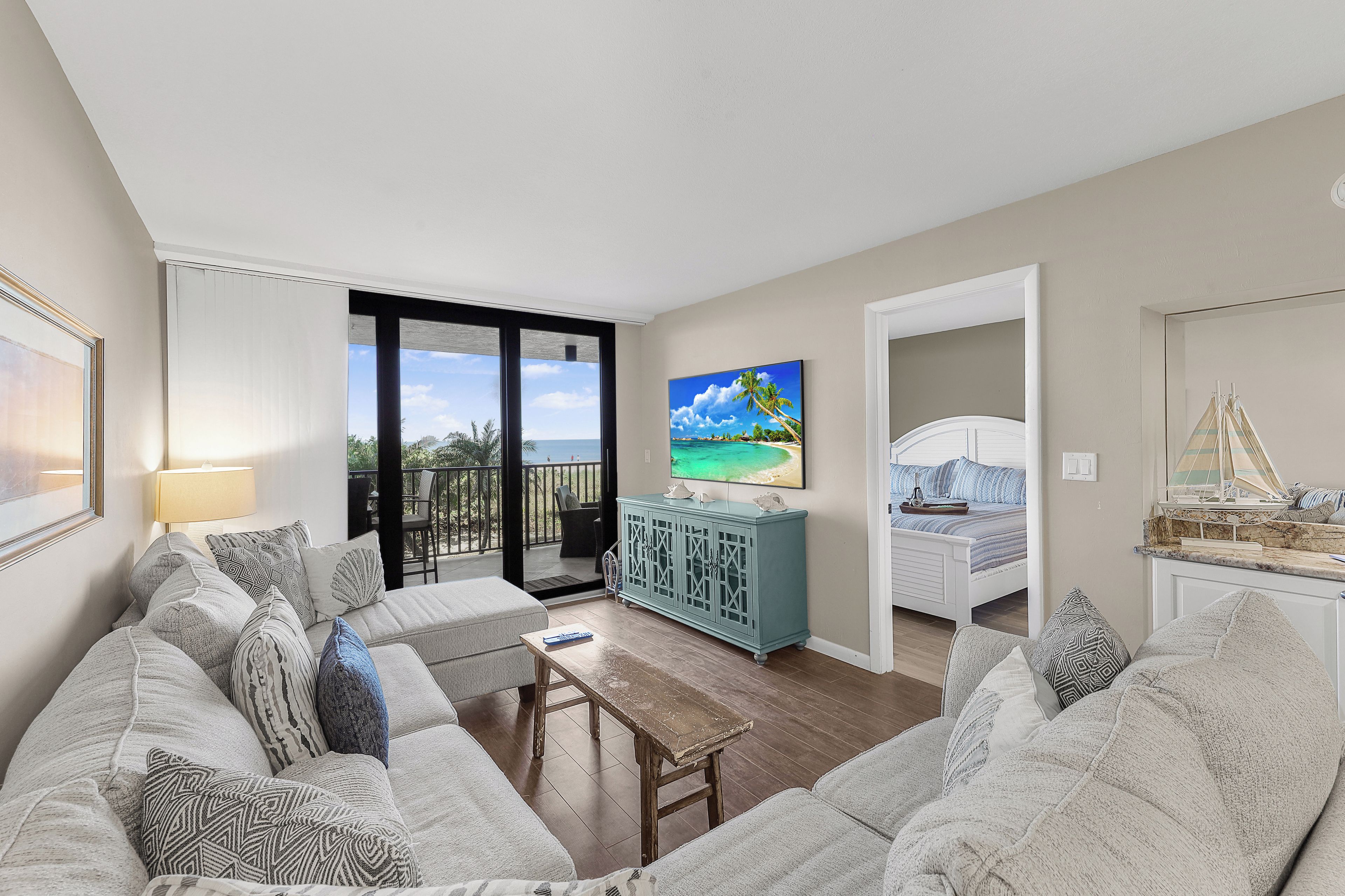 A living room with a sofa, a coffee table, and a TV. There is a balcony with a view of the ocean.