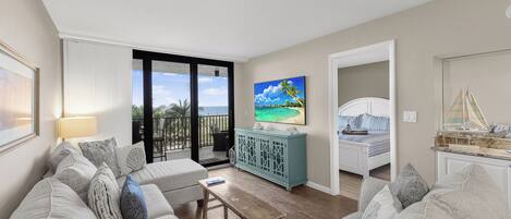 A living room with a sofa, a coffee table, and a TV. There is a balcony with a view of the ocean.