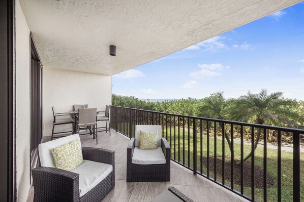 A balcony with wicker furniture, a table, and chairs, overlooking a lush garden and distant landscape.