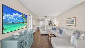 Een ruime woonkamer met een grote flatscreen televisie, een lichtgekleurde bank en een houten salontafel.